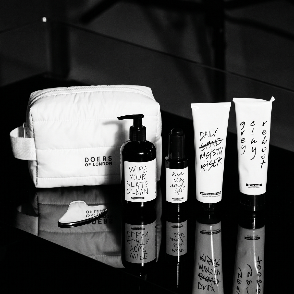 Black and white scene of the Looksmaxxing Kit: the white Doers toiletry bag with cleanser pump, clear serum bottle, hydrating face cream tube, detox mask tube and gua sha tool on a reflective black surface.