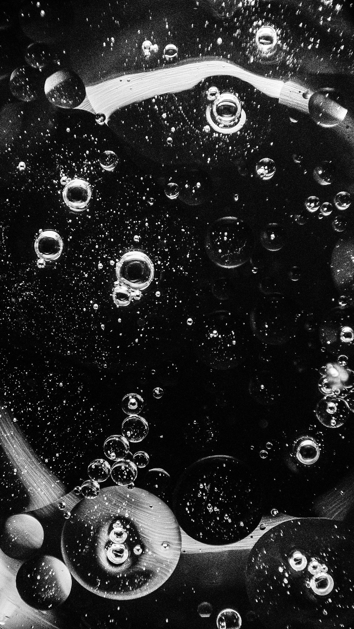 Macro black and white shot of oil droplets and bubbles suspended in water against a dark background.