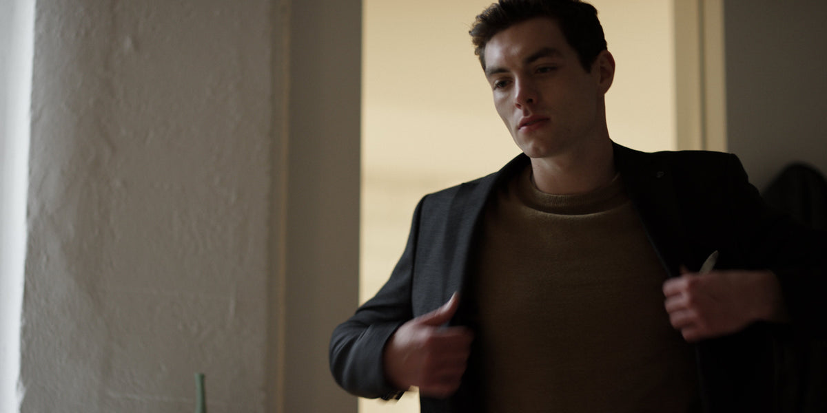 A young man adjusts his open blazer over a knit sweater in warm window light beside a textured white wall.