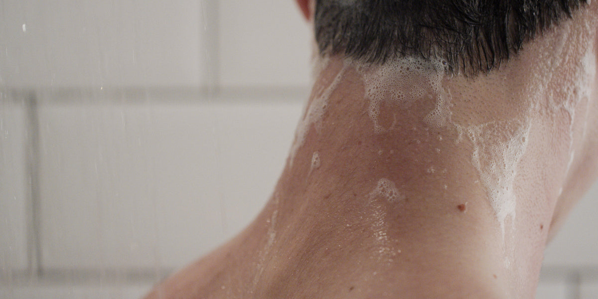 Close-up of a man's neck and hairline in the shower with shampoo suds running down over white tiles.