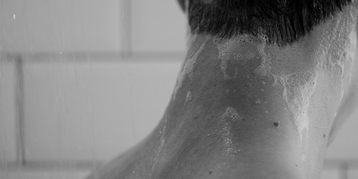 Black and white close-up of a man's neck and hairline in the shower with shampoo suds running down his back.