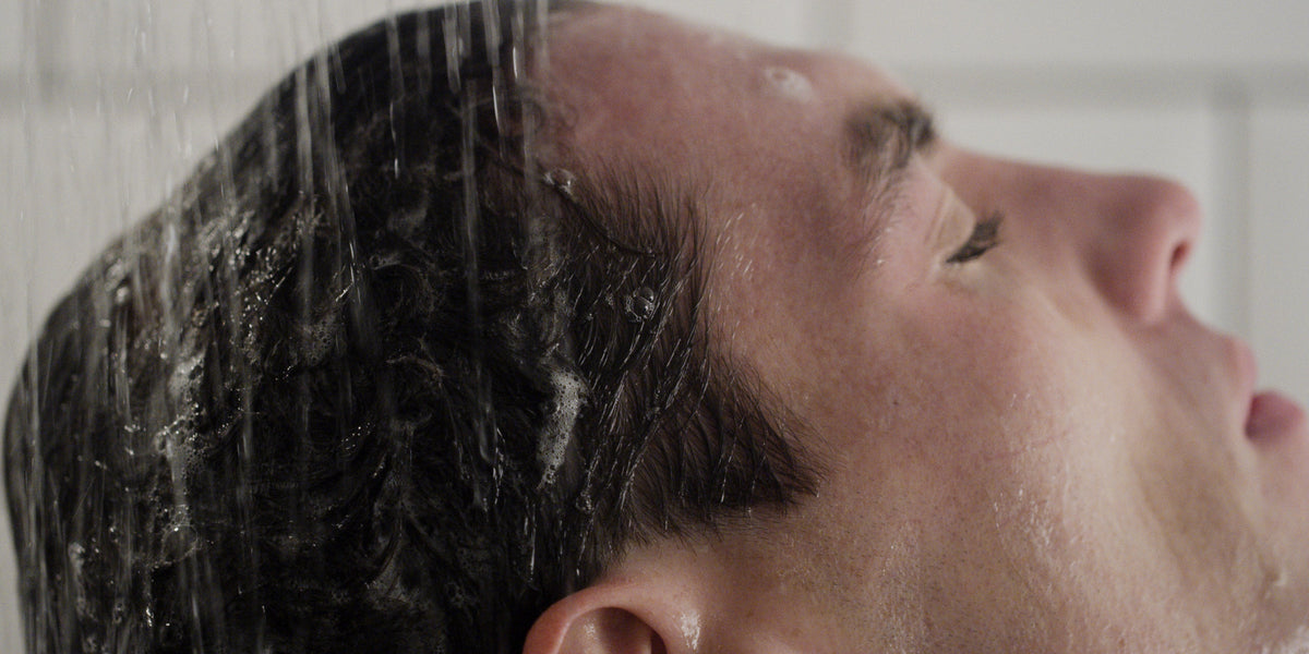 Side profile of a man with water streaming through his hair under the shower, eyes closed.