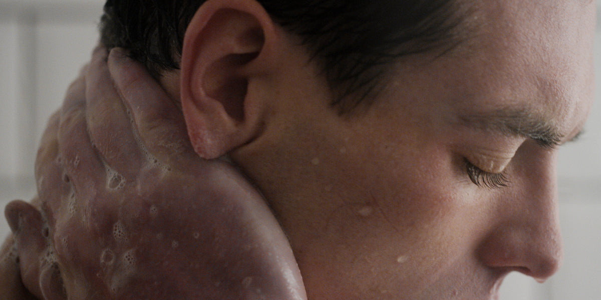 Close-up of a man's hand pressing foamy cleanser to his cheek and temple, eyes closed.