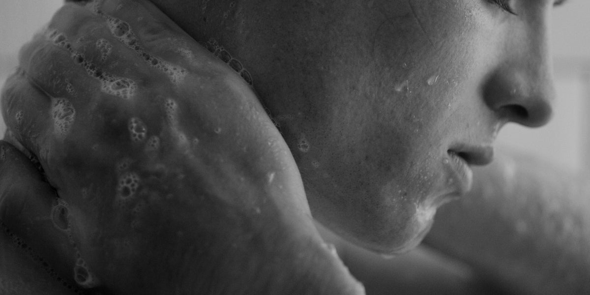 Black and white close-up of a foamy hand pressing cleanser against a man's jaw and neck.