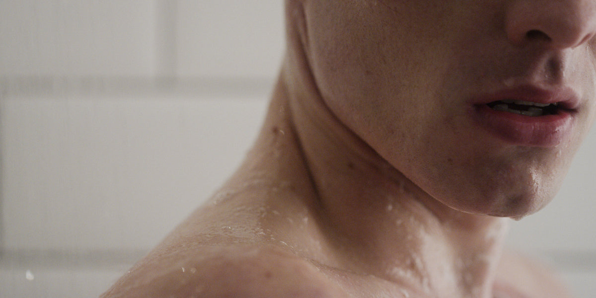 Tight close-up of a man's wet jaw, neck and shoulder in the shower with white tile behind.