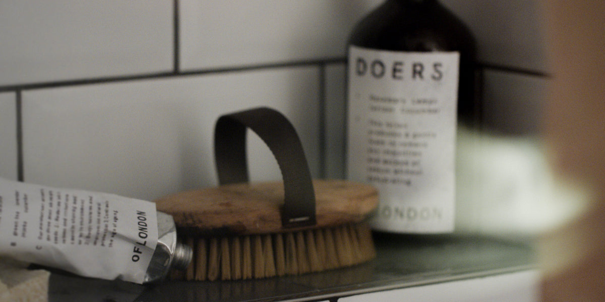 Still life of a Doers tube, a wooden body brush and a Doers bottle on a bathroom shelf against white tile.