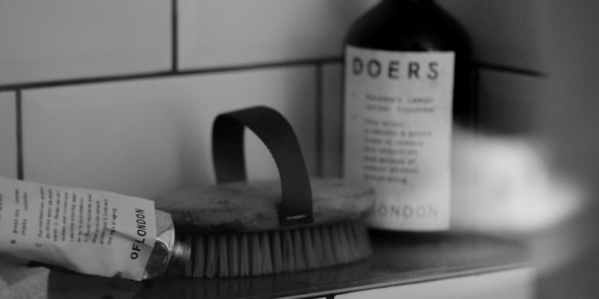 Black and white still life of a Doers tube, body brush and bottle on a bathroom shelf against tile.