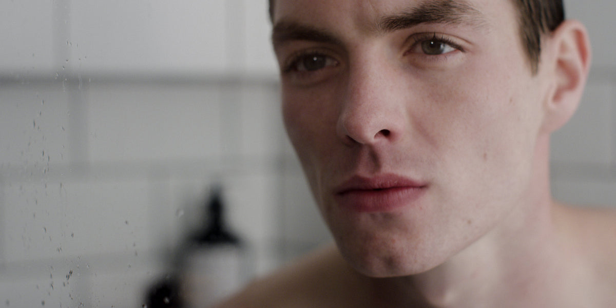 Close-up portrait of a young man in a tiled bathroom, looking at the camera with light stubble.