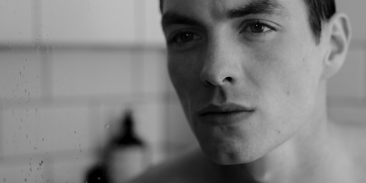 Black and white close-up portrait of a young man in a tiled bathroom looking at the camera.