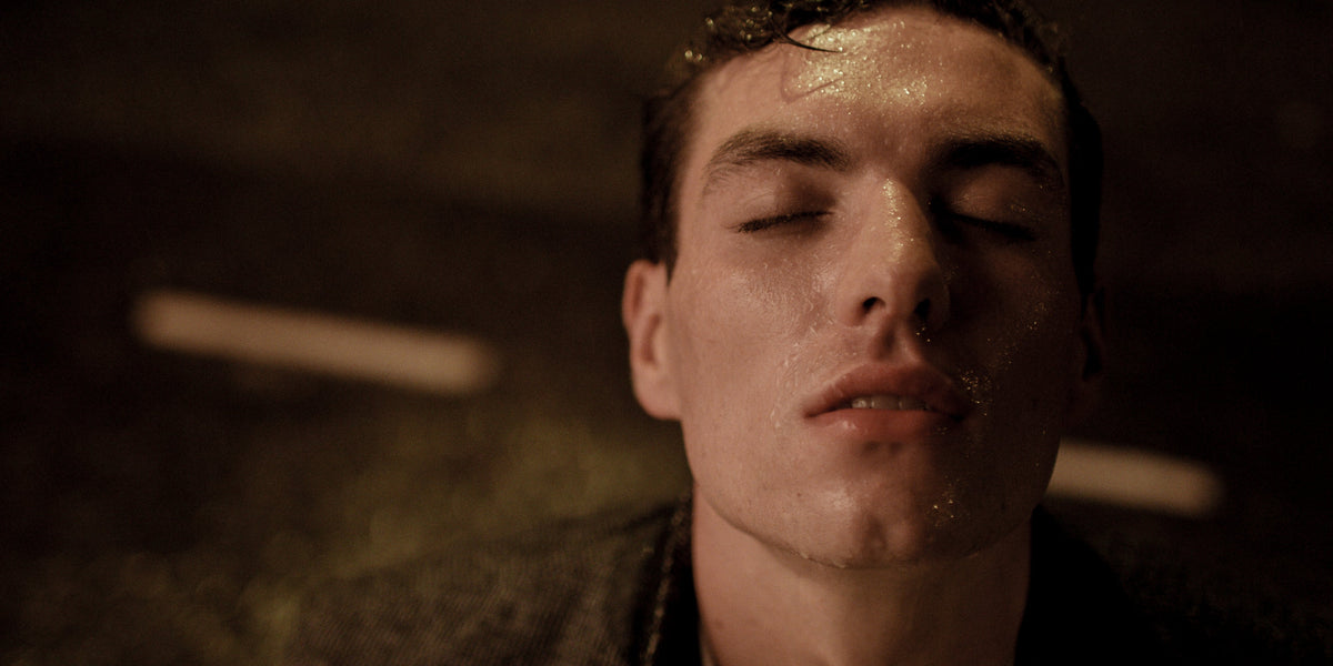 Close-up of a man's face glistening with water, eyes closed, lit warmly with blurred lights behind.