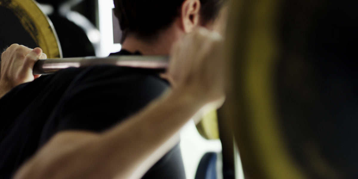 Man performing a barbell back squat in a gym, photographed from behind with weight plates in frame.