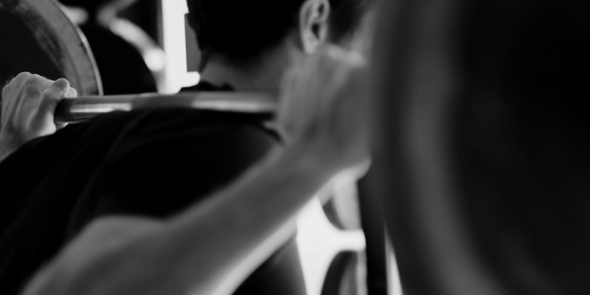 Black and white shot of a man performing a barbell back squat in a gym, photographed from behind.