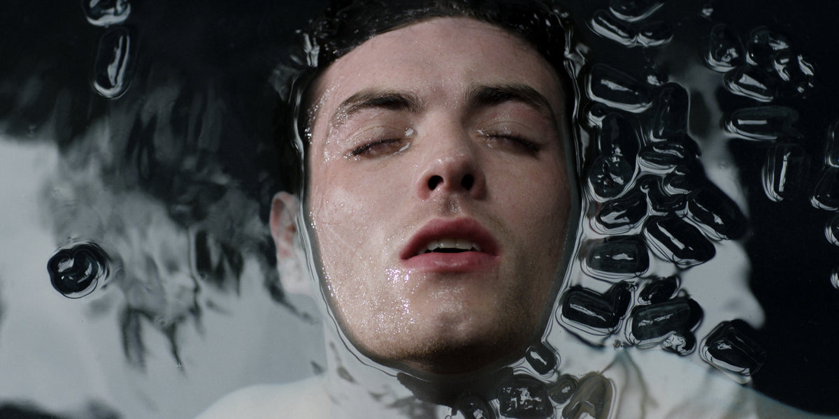 Close-up of a man's face surfacing in water with mouth open, droplets on skin and ice cubes drifting.
