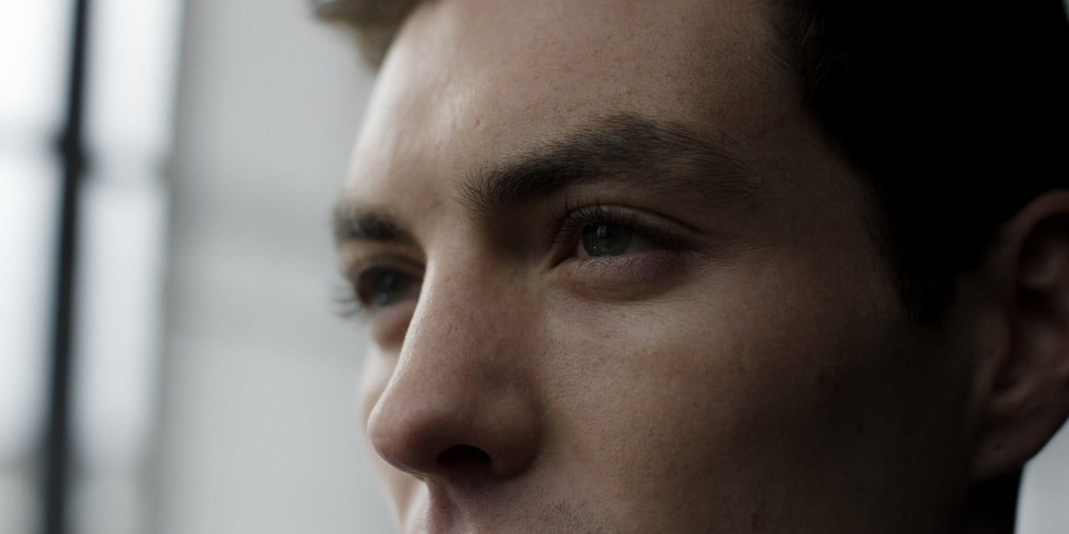 Three-quarter close-up of a young man's eye, brow and cheek with soft window light revealing freckles and stubble.