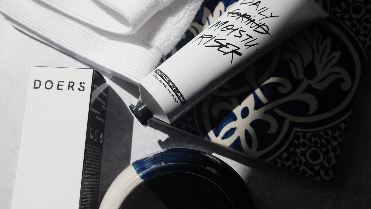 Hydrating Face Cream tube with handwritten Daily Grind Moisturiser text, alongside the Doers box, towel and ceramic dish on patterned blue tile.