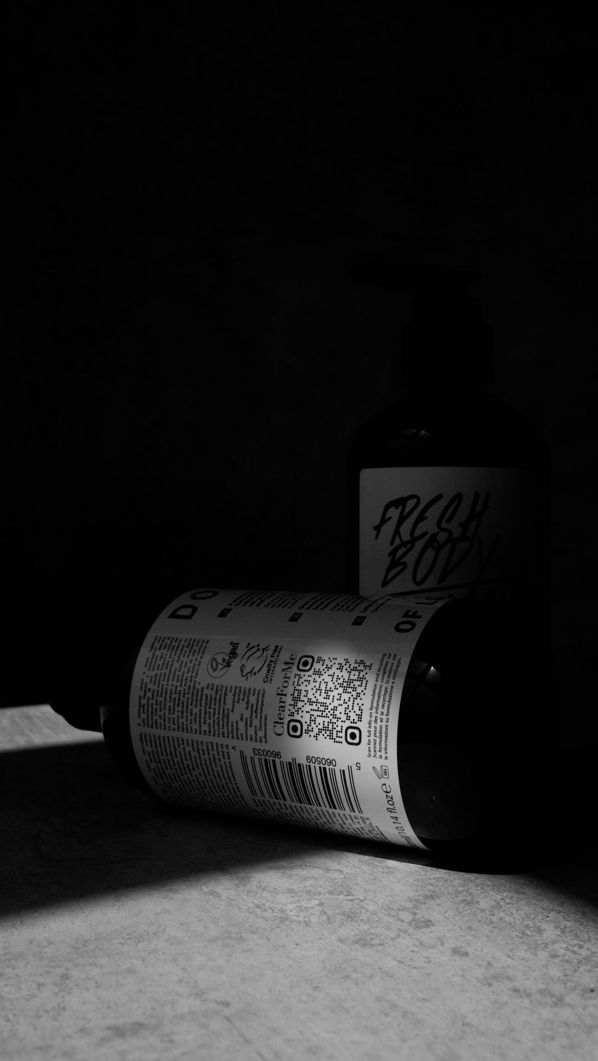 Black and white moody shot of a Hand & Body Wash bottle upright behind a second bottle tipped on its side showing the ingredients label.