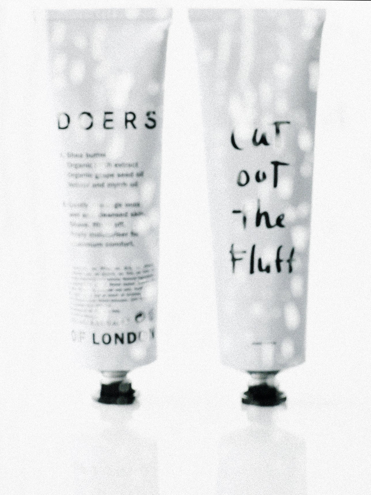 Facial cleanser tube and shave cream Cut Out the Fluff tube side by side seen through a glass screen with bokeh highlights.