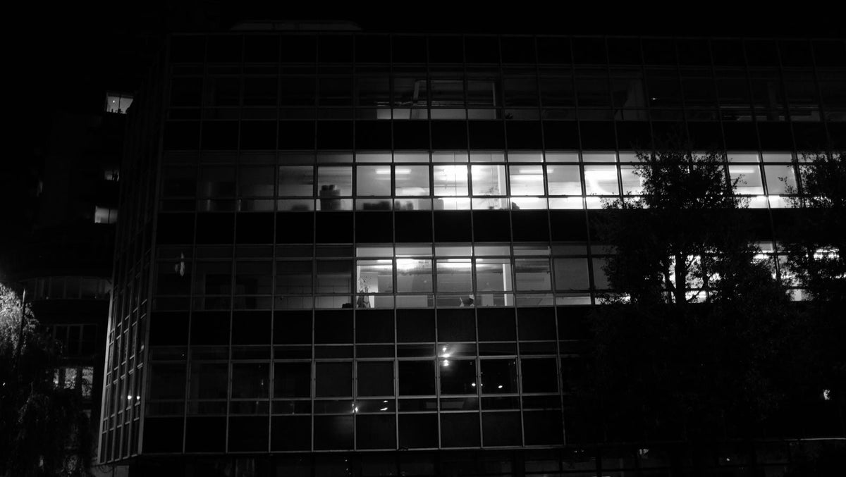 Black and white shot of a modernist glass office facade at night with two floors of fluorescent-lit interiors.