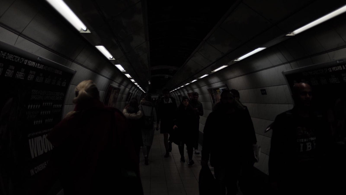 Commuters walk through a tiled London Underground passageway in moody low light, film posters on the walls.