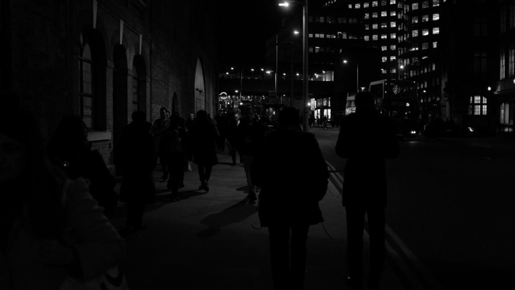 Black and white shot of pedestrians on a city pavement at night with lit office buildings in the background.