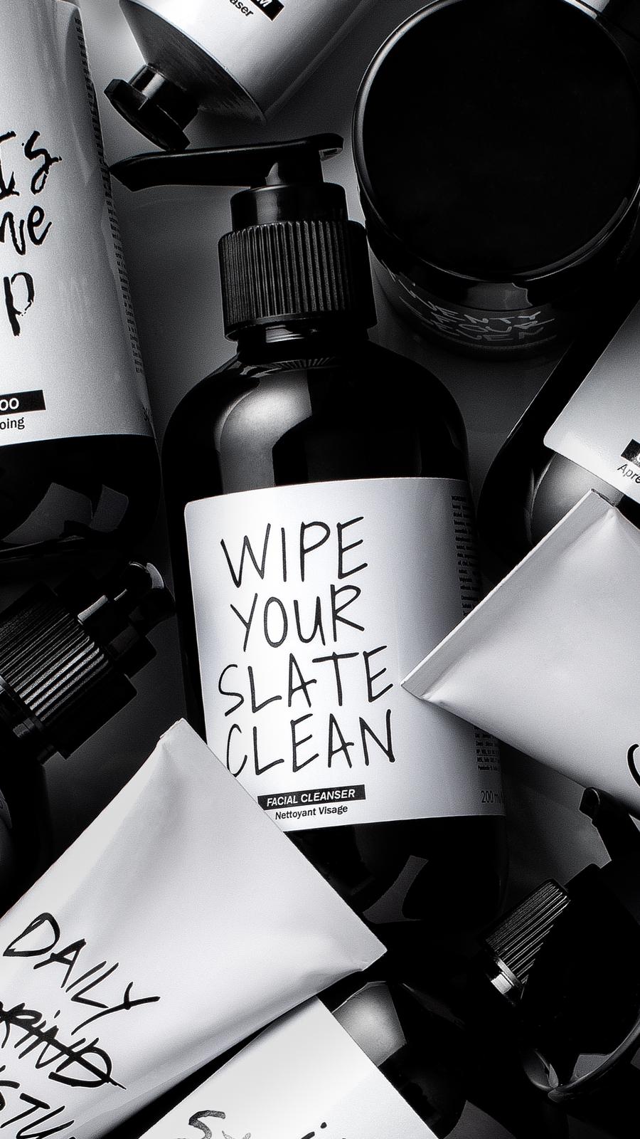 Black and white close-up of the facial cleanser pump bottle (Wipe Your Slate Clean) surrounded by other range pieces.